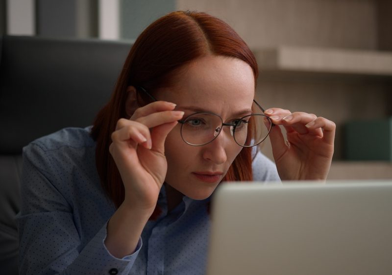 Woman wearing and adjusting bifocal eyeglasses while looking at a laptop screen, illustrating how bifocals support near and distance vision - bifocals meaning