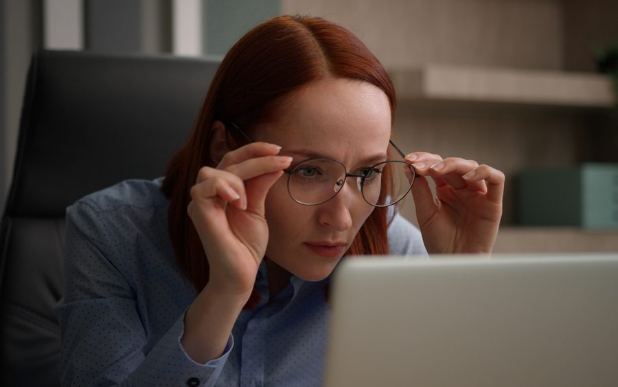 Woman wearing and adjusting bifocal eyeglasses while looking at a laptop screen, illustrating how bifocals support near and distance vision - bifocals meaning