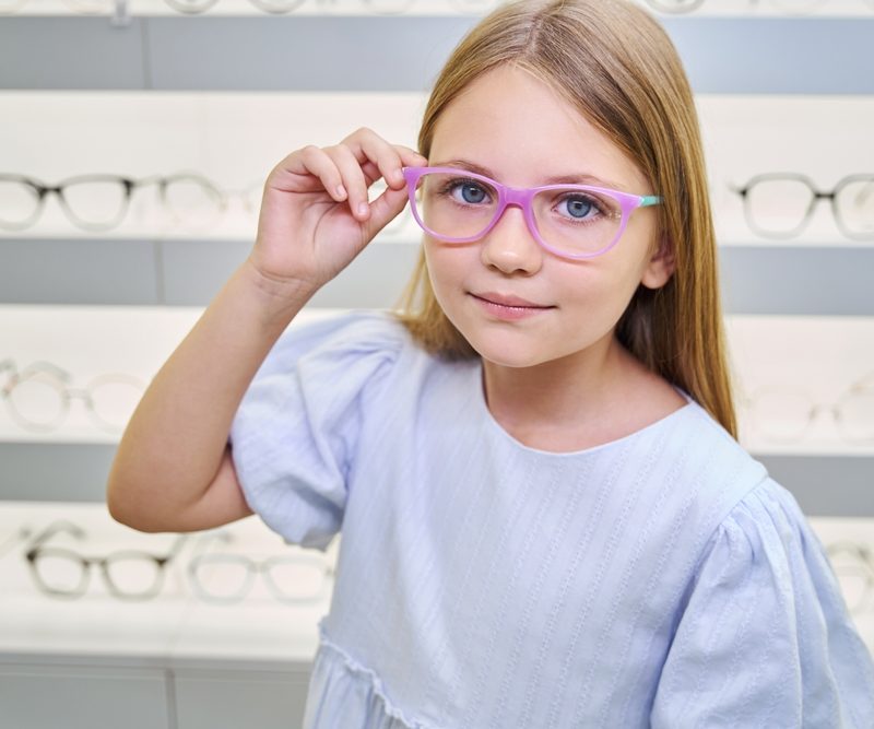 Smiling young girl wearing eyeglasses inside an optical store, representing a routine pediatric vision screening - children's eye test