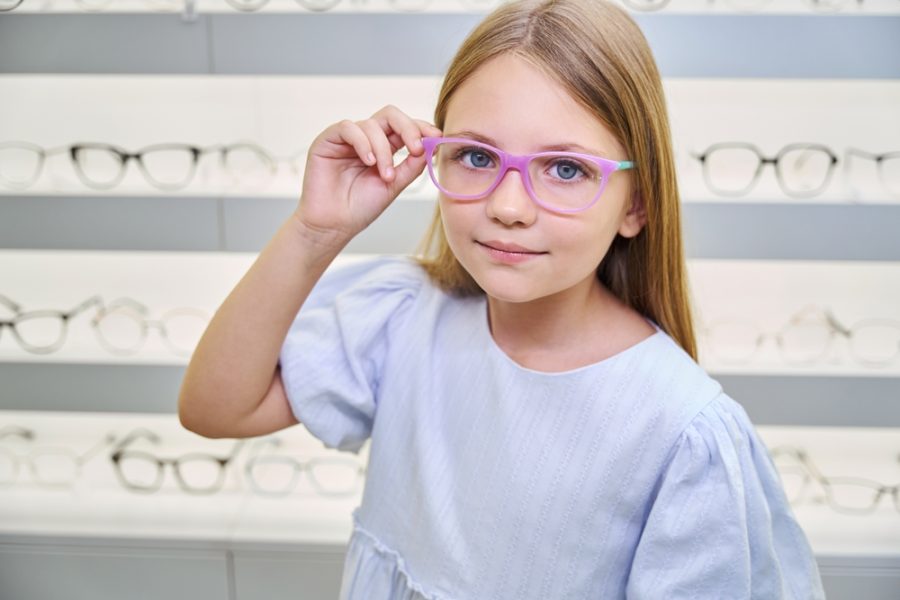 Smiling young girl wearing eyeglasses inside an optical store, representing a routine pediatric vision screening - children's eye test