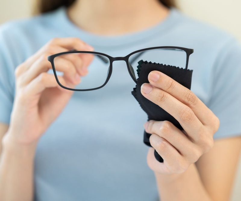 Person holding eyeglasses and a microfiber cloth while cleaning lenses with spray solution - glass cleaner on glasses
